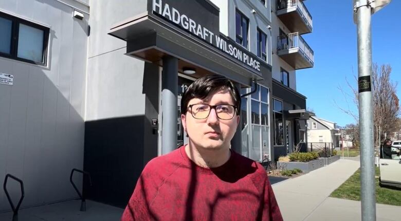 A man in glasses and a red shirt stands in front of a building that reads Hadgraft Wilson Place. 