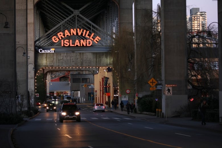 A photo of a large red neon sign reading 'Granville Island' hangs over the entrance to a market underneath a bridge.