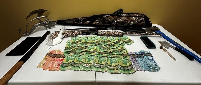 Cash, two guns, a white bag of powder, axes and more are laid out on a white table. 