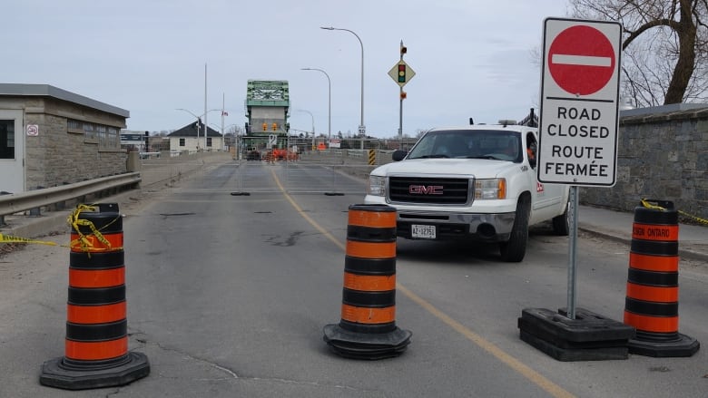 A white pickup truck sits behind a row of pylons and a road closed sign. In the background a green bridge can be seen, blocked off by fencing.