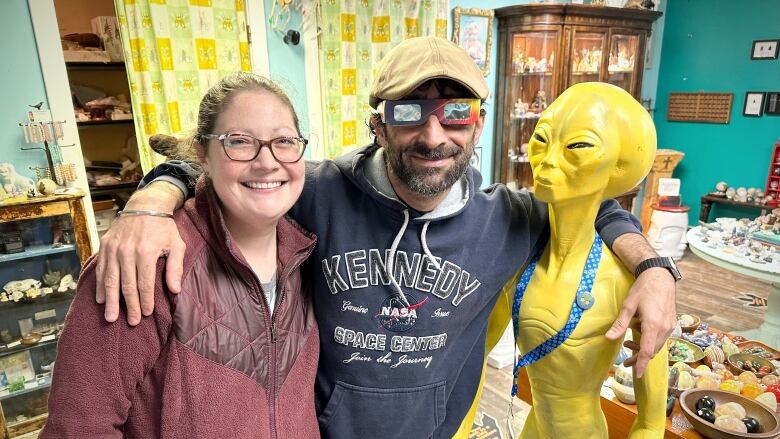 A man with a blue sweatshirt and tan cap wears shiny dark glasses with his arms around a woman in a red coat and glasses and a model of a green alien.