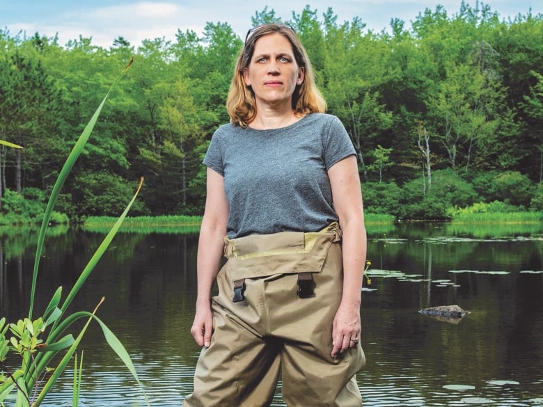 Woman wearing waist high waders stands in a pond. She is looking straight into the camera.