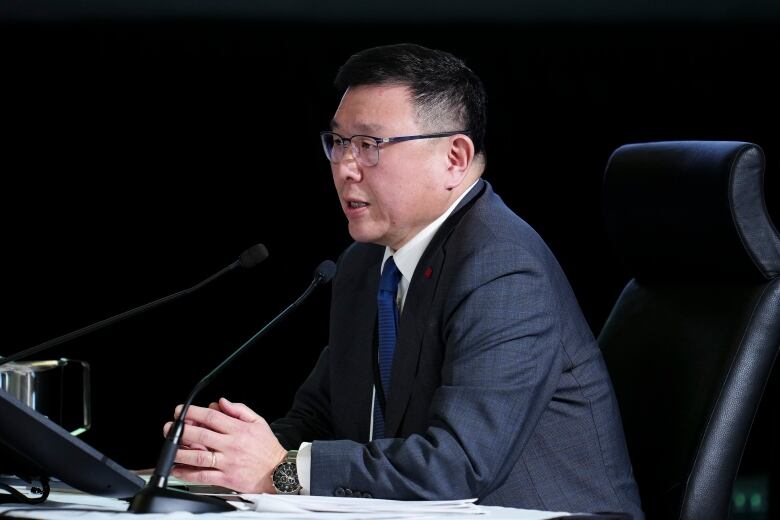 A man wearing a suit sits at a table speaking into a conference microphone