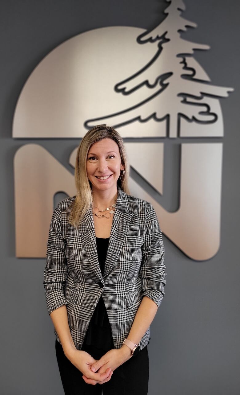 A woman with blonde hair and a checkered blazer standing in front of a grey wall with a large metal sign. 