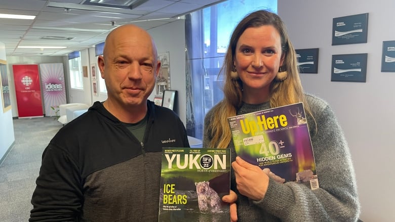 A man and woman stand in a hallway, holding up 2 magazines.