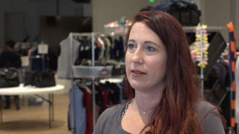 A woman stands in front of clothing racks. She's wearing a grey shirt and silver necklace. 