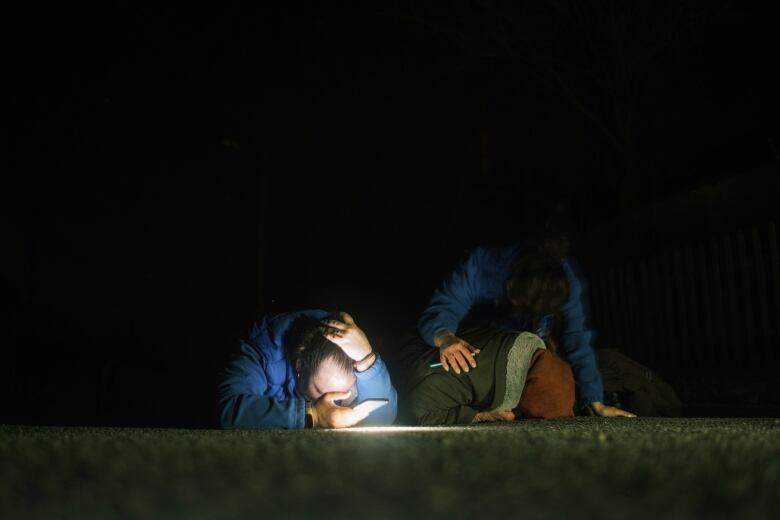 Three people lie face down on the pavement in the darkness. One man covers his head with his hand.