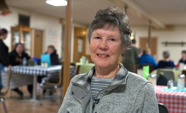 A woman with short grey hair and a grey sweater smiles while seated at a table inside a recreation centre.