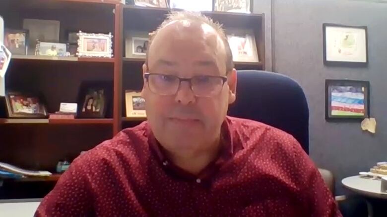 A man with glasses and a red button-up shirt sits in an office with a large bookshelf behind him. 