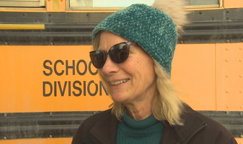 A woman with blonde hair and a teal toque stands in front of a school bus smilng.