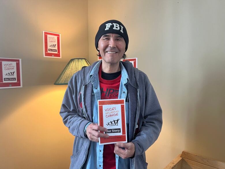 A man in a toque and grey jacket holds a copy of a book titled Voices Influencing Change: Our Story