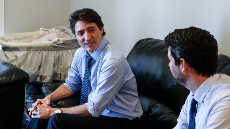 A man sits on a black couch, speaking to another man on the couch.