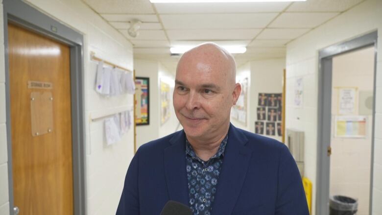 Photograph of a man in a blue suit talking into a microphone in a corridor. 