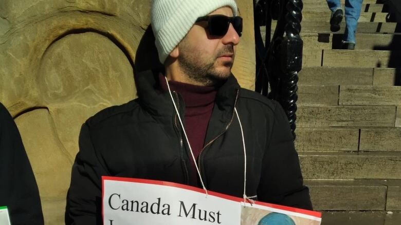 A man with sunglasses hold a sign at a protest in Ottawa asking for family members in Gaza to be admitted to Canada.