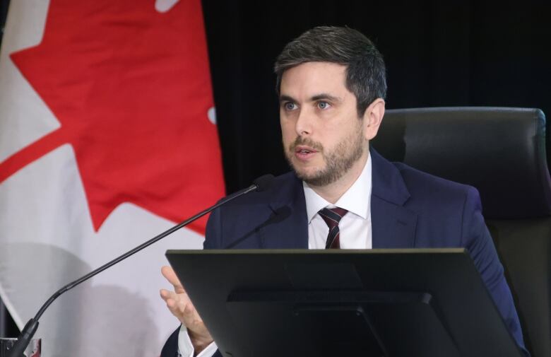 A man in a dark blue suit and tie sits at a table and speaks into a microphone. A Canadian flag is drapped in the background.