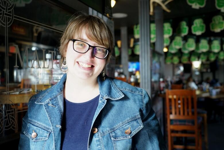 A woman wearing glasses and jean jacket stands in a pub.