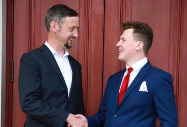 Two smiling men, one middle aged and one younger, shake hands. They're wearing suits.