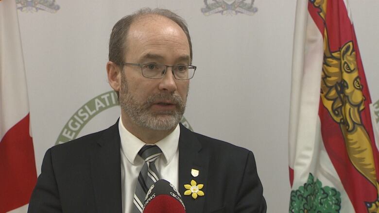 A man with glasses and a greying beard dressed in a suit speaks into a microphone with a PEI flag in the background.