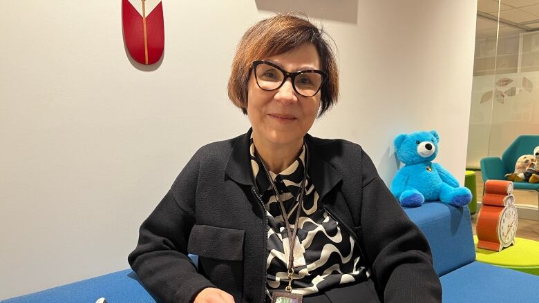 A woman sits on a couch, with a blue teddy bear in the background.