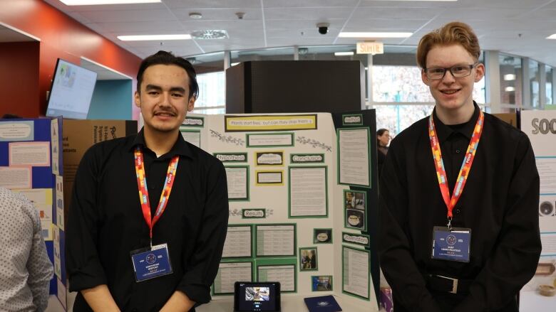 Two teenage boys wearing nametags on lanyards pose beside a bristol-board display.