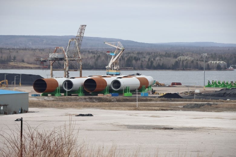 Several large grey-and-rust-coloured steel tubes lie on their sides on a cleared piece of land on a pier next to water.