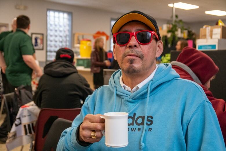 A person wearing a baseball cap, red sunglasses and a blue hoodie holds a coffee mug in a crowded room.