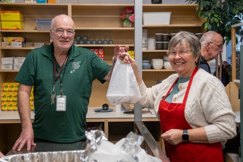 Two people stand in a kitchen-like setting, holding a white plastic bag between them. They are smiling.