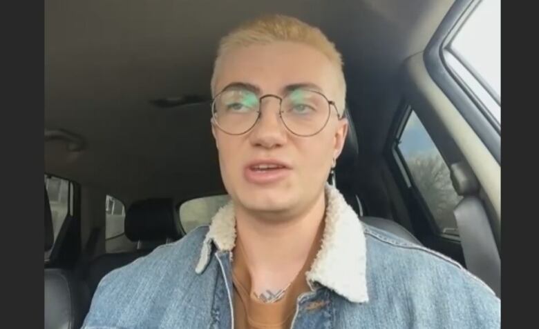 A person with blonde hair and a jean jacket sits in their car.