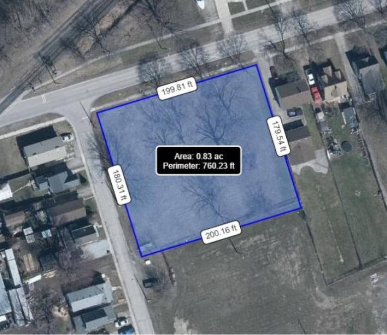 An aerial shot of an empty plot of land. To the left and top of the lot are an intersecting roadway. Houses sit along the left side of the road opposite the empty lot. Three houses sit on the properties to the right of the lot.