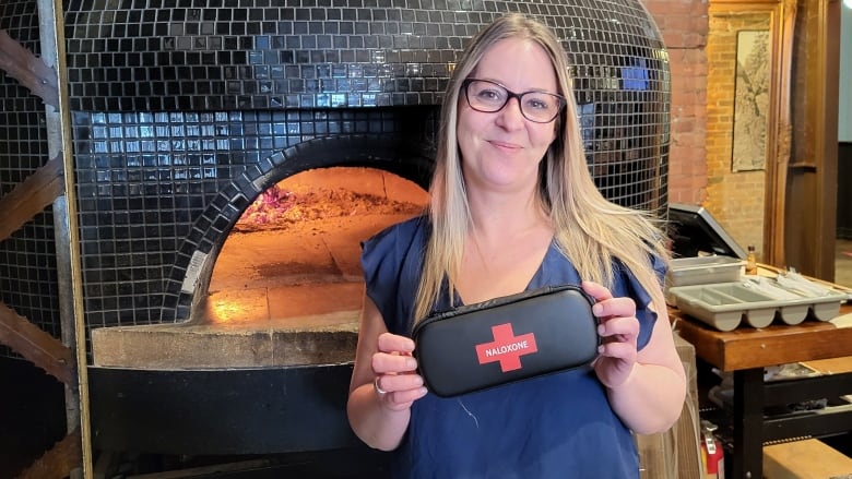 An adult woman wearing a blue shirt holds a black pouch with a red cross on it, standing in front of a wood and brick pizza over inside a restaurant.