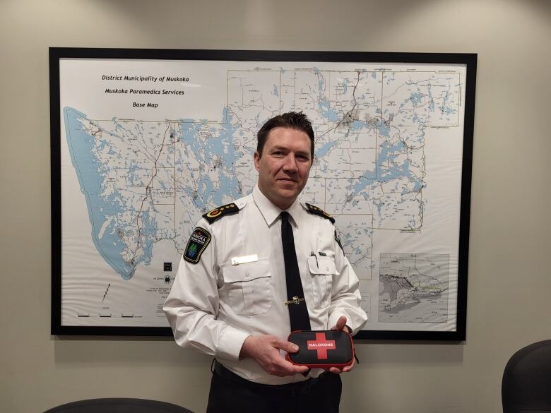 A caucasian male in a white first responders' shirt with medical service logos holds a black pouch with a red cross on its front stands in front of a topographic map of a region in Ontario, inside an office space.