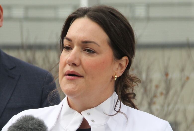 A woman in a white medical or lab coat stands at a podium, speaking into a microphone.