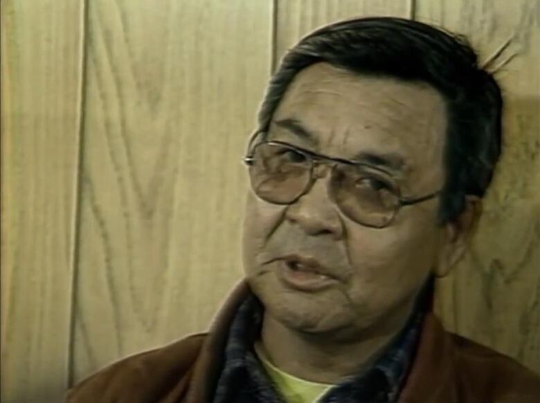 A man speaks against a backdrop of 70s era wood panelling. 