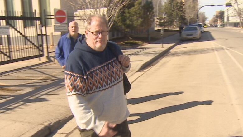 A man is crossing the street, while wearing a hooded sweater and glasses, with a bag on his shoulder.