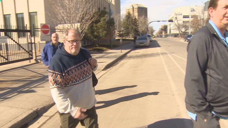 A man is crossing the street, while wearing a hooded sweater and glasses, with a bag on his shoulder.