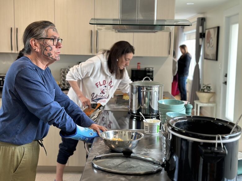 A bespectacled man with long grey hair pulled back in a ponytail and wearing a blue sweater stands at a cluttered kitchen counter, looking out a window. There is a brown-haired woman behind him.