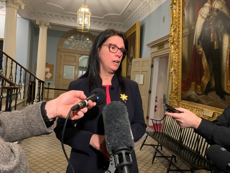 A woman with dark hair and glasses speaks to reporters holding microphones.