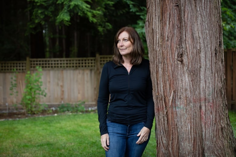 A white woman with black hair leans against a tree.