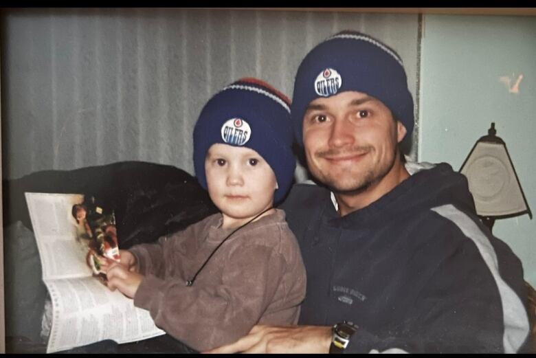 A man smiles at the camera, with a child on his lap. Both are wearing dark blue tuques with an Edmonton Oilers patch on the front.