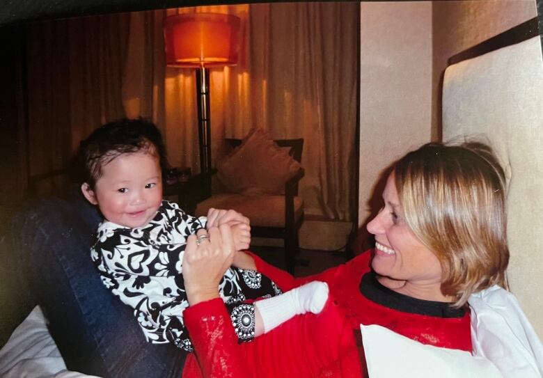 A toddler gives a big smile to the photographer while sitting on the lap of a woman who is smiling at the child.