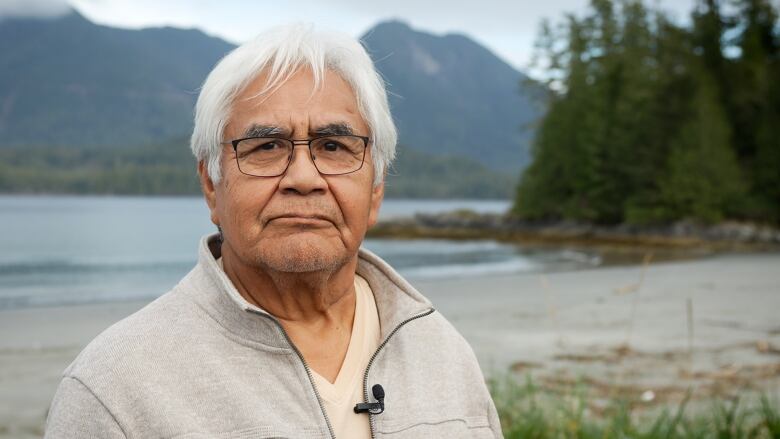 A man looks at the camera standing near the beach with white hair and a tanned jacket.  