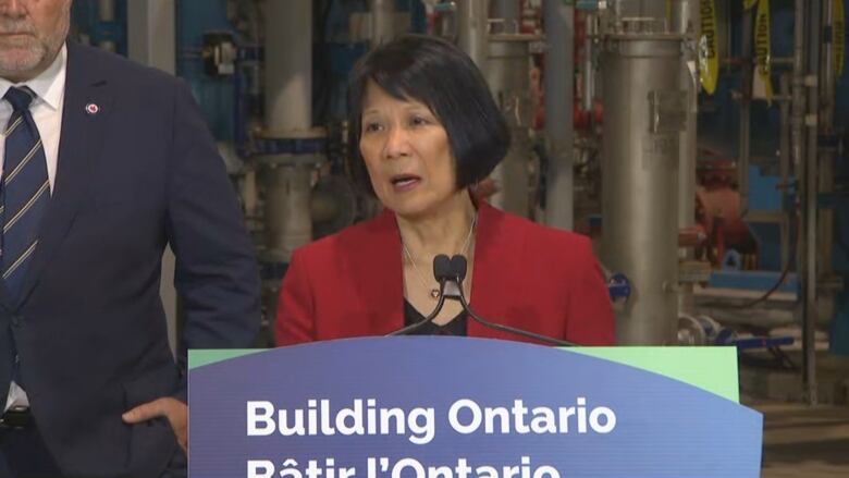 A middle-aged woman speaks into a microphone at a podium indoors. A man is standing behind her in a suit. There are pipes behind her also.