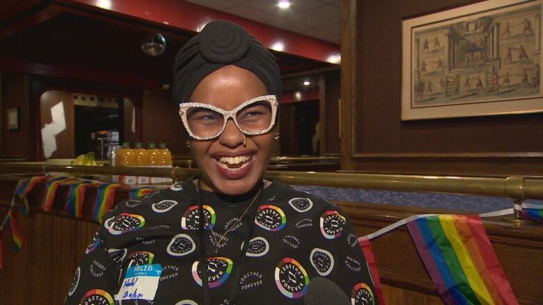 Woman in sparkly glasses and rainbow decorated shirt stands in bar smiling joyously.  