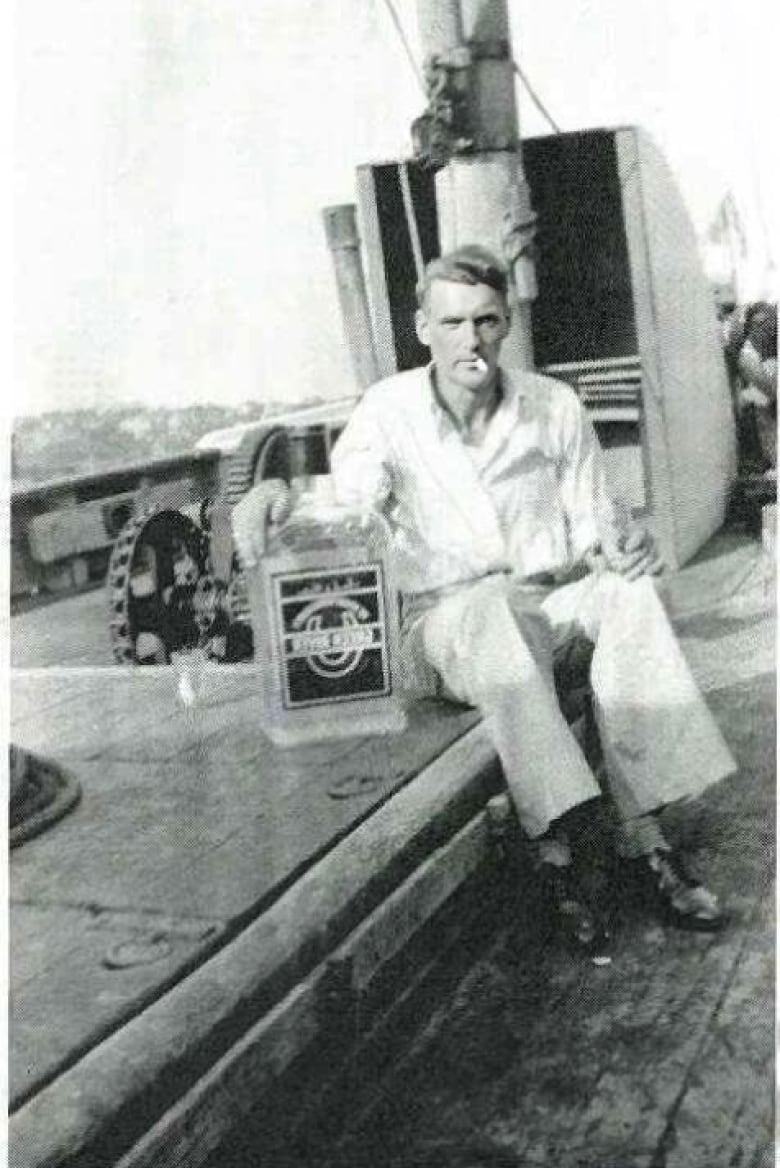 Black and white photo of a man sitting on a boat deck next to a very large glass container of alcohol.