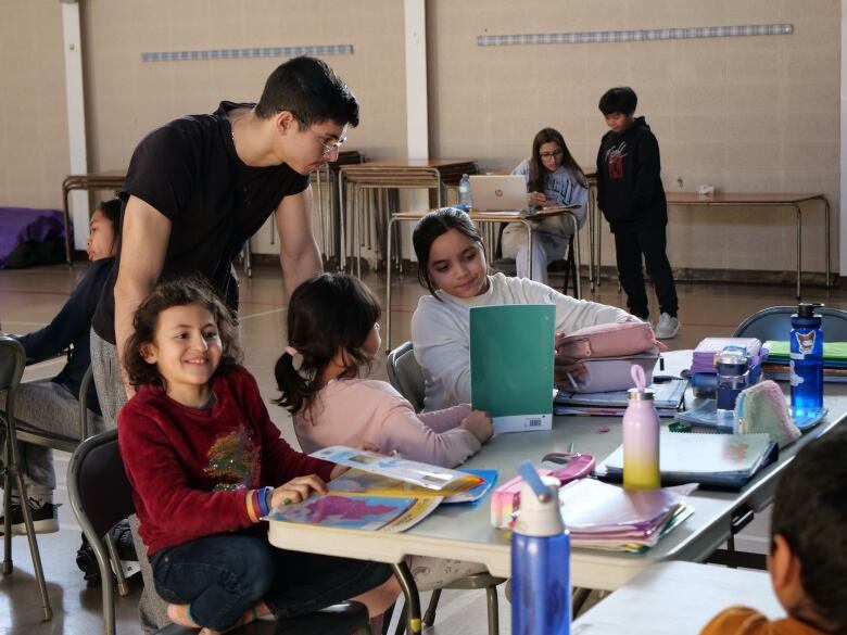 Young children in a classroom sitting at a long table with school books laid out in front of them. 