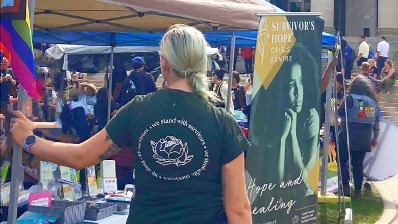 A photo of a woman outdoors in front of a stand with a banner that reads 'Survivor's Hope Crisis Centre.'