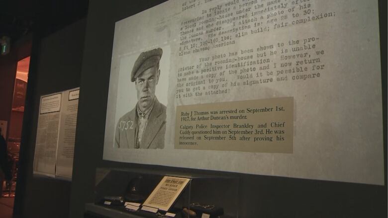 A slideshow is projected on a wall above a glass case of artifacts in a museum. the image on the wall is a mug shot of a man in black and white.