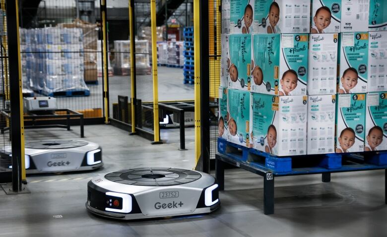 a cylindrical silver robotic device sits beside a pallet of diaper boxes in a warehouse