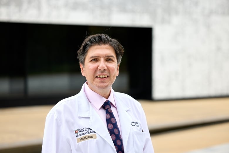 A man with silver-black hair smiles at the camera, he is wearing a pink shirt and dark blue tie with a white medical jacket overtop.