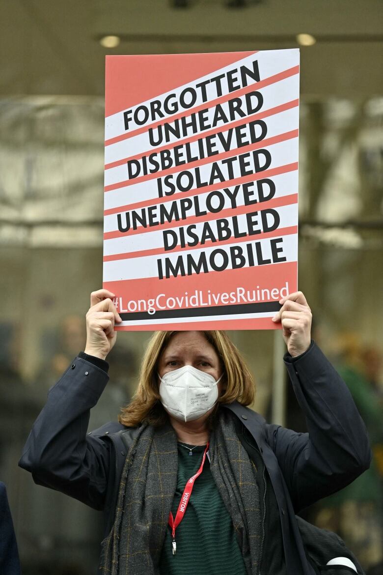 A woman faces the camera, she wears a facemask. She's holding up a sign that says 